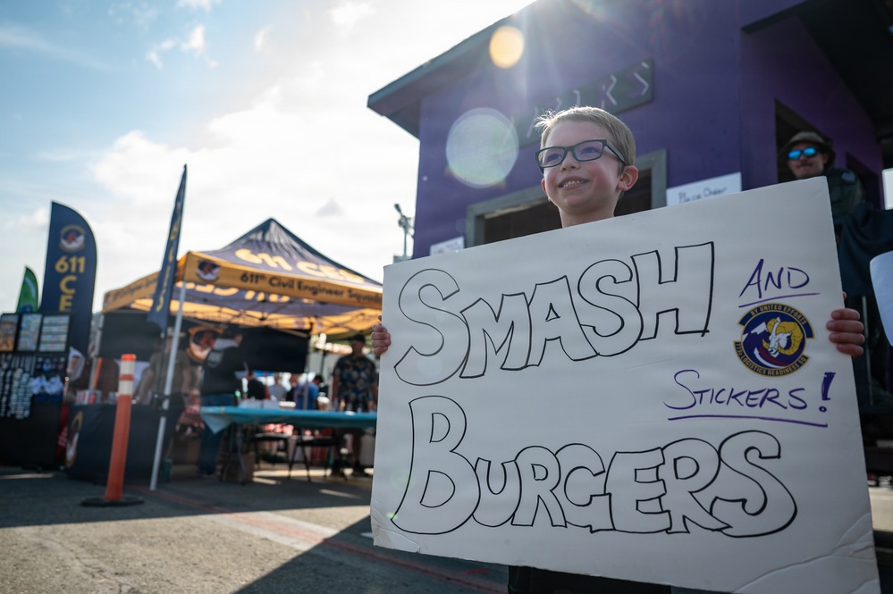 A young child wearing glasses holds a handmade white sign reading 'SMASH BURGERS AND STICKERS!' at what appears to be an outdoor military community event. In the background, a yellow canopy tent displays '611th Civil Engineer Squadron' branding, along with a purple building and various event booths. The event appears to be associated with the U.S. Air Force, as indicated by military unit insignia visible on the sign and banners.