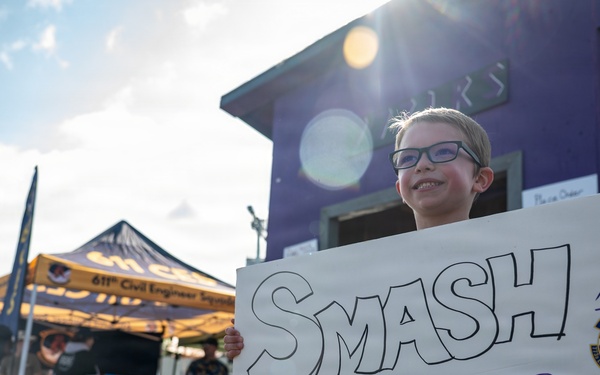 A young child wearing glasses holds a handmade white sign reading 'SMASH BURGERS AND STICKERS!' at what appears to be an outdoor military community event. In the background, a yellow canopy tent displays '611th Civil Engineer Squadron' branding, along with a purple building and various event booths. The event appears to be associated with the U.S. Air Force, as indicated by military unit insignia visible on the sign and banners.