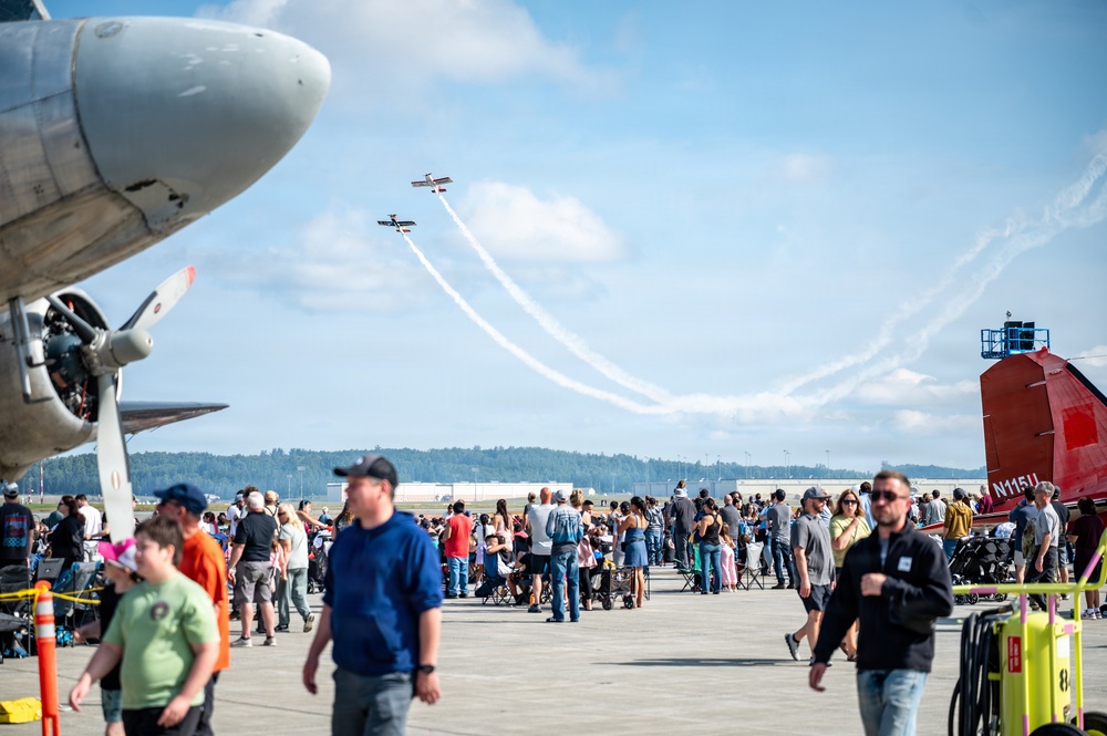 An outdoor airshow event at an airport tarmac with a large crowd of spectators watching two small aerobatic aircraft performing crossing maneuvers with smoke trails in a partly cloudy sky. A large propeller-driven aircraft nose is visible in the foreground left, and a red aircraft tail with registration N115U is visible on the right. The event appears well-attended with hundreds of people gathered on the tarmac.