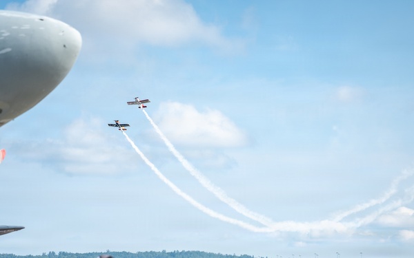 An outdoor airshow event at an airport tarmac with a large crowd of spectators watching two small aerobatic aircraft performing crossing maneuvers with smoke trails in a partly cloudy sky. A large propeller-driven aircraft nose is visible in the foreground left, and a red aircraft tail with registration N115U is visible on the right. The event appears well-attended with hundreds of people gathered on the tarmac.