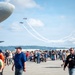 An outdoor airshow event at an airport tarmac with a large crowd of spectators watching two small aerobatic aircraft performing crossing maneuvers with smoke trails in a partly cloudy sky. A large propeller-driven aircraft nose is visible in the foreground left, and a red aircraft tail with registration N115U is visible on the right. The event appears well-attended with hundreds of people gathered on the tarmac.