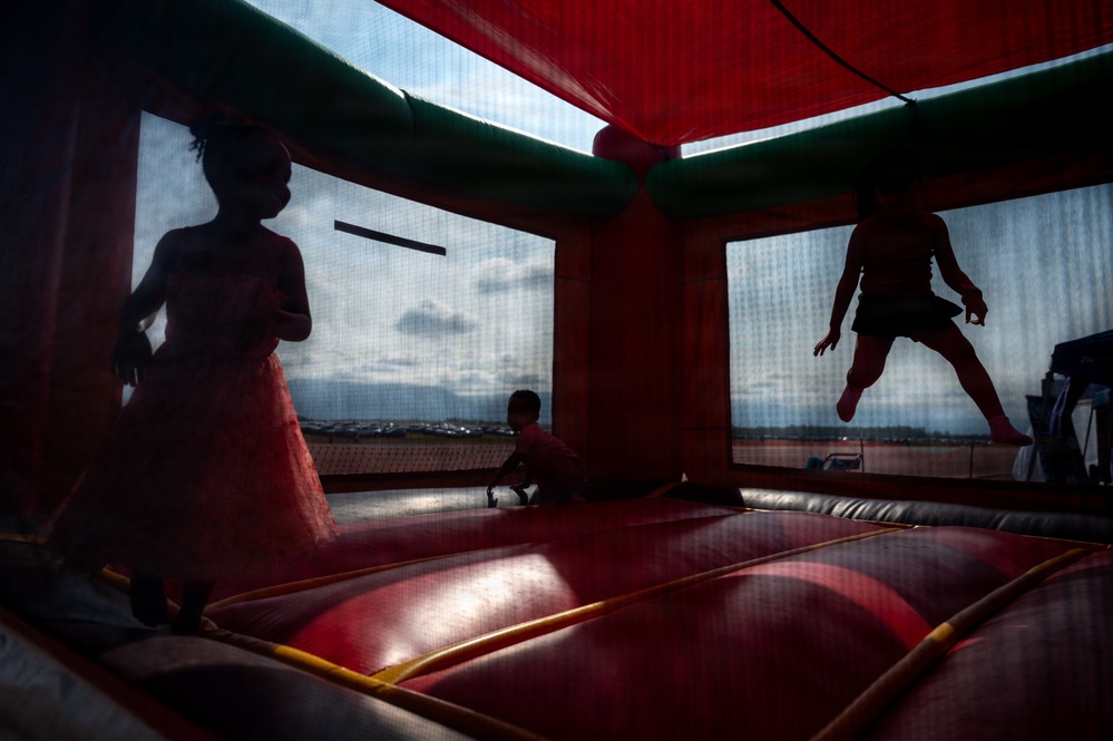 Three children are playing inside a red and green inflatable bounce house. The image is taken from a low angle inside the structure, creating dramatic silhouettes of the children against the mesh windows. One child appears to be mid-jump, one is standing in a dress, and one is crouching on the inflatable floor.