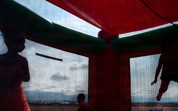 Three children are playing inside a red and green inflatable bounce house. The image is taken from a low angle inside the structure, creating dramatic silhouettes of the children against the mesh windows. One child appears to be mid-jump, one is standing in a dress, and one is crouching on the inflatable floor.