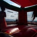 Three children are playing inside a red and green inflatable bounce house. The image is taken from a low angle inside the structure, creating dramatic silhouettes of the children against the mesh windows. One child appears to be mid-jump, one is standing in a dress, and one is crouching on the inflatable floor.