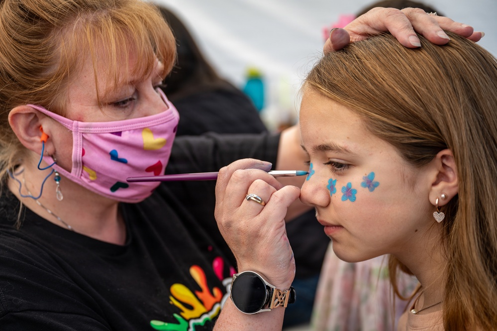 A face painter wearing a pink patterned mask is applying blue flower designs to a young girl's cheek using a fine brush. The artist steadies the girl's head with one hand while carefully painting near her nose. Three small blue flower designs are already visible on the girl's cheek.