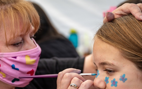 A face painter wearing a pink patterned mask is applying blue flower designs to a young girl's cheek using a fine brush. The artist steadies the girl's head with one hand while carefully painting near her nose. Three small blue flower designs are already visible on the girl's cheek.