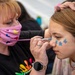 A face painter wearing a pink patterned mask is applying blue flower designs to a young girl's cheek using a fine brush. The artist steadies the girl's head with one hand while carefully painting near her nose. Three small blue flower designs are already visible on the girl's cheek.