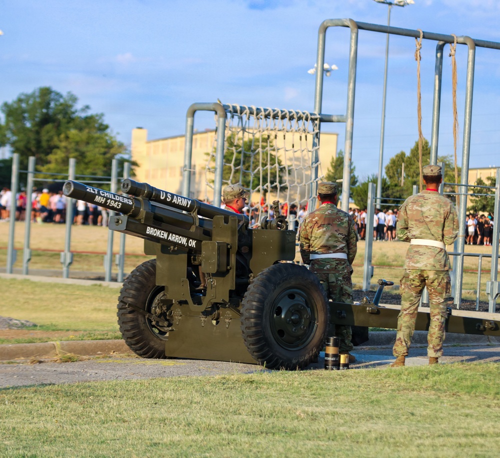 A U.S. Army ceremonial cannon is positioned on a military installation grounds, with three soldiers in MultiCam/OCP uniforms standing nearby. The cannon bears inscriptions reading 'U.S. ARMY', '2LT CHILDERS MH 1943', and 'BROKEN ARROW, OK', indicating it is a named/dedicated historical piece. A large group of people, likely trainees or recruits, can be seen assembled in the background near what appears to be an obstacle course.