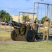 A U.S. Army ceremonial cannon is positioned on a military installation grounds, with three soldiers in MultiCam/OCP uniforms standing nearby. The cannon bears inscriptions reading 'U.S. ARMY', '2LT CHILDERS MH 1943', and 'BROKEN ARROW, OK', indicating it is a named/dedicated historical piece. A large group of people, likely trainees or recruits, can be seen assembled in the background near what appears to be an obstacle course.