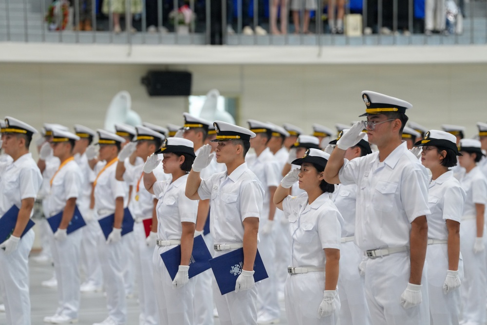 A formal military or naval ceremony featuring personnel in white dress uniforms executing a salute in organized formation. The individuals are wearing white caps with gold/black bands, white uniforms, white gloves, and white belts, consistent with naval dress attire. Some personnel are holding blue folders or documents, suggesting this may be a graduation or commissioning ceremony.