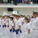 A formal military or naval ceremony featuring personnel in white dress uniforms executing a salute in organized formation. The individuals are wearing white caps with gold/black bands, white uniforms, white gloves, and white belts, consistent with naval dress attire. Some personnel are holding blue folders or documents, suggesting this may be a graduation or commissioning ceremony.