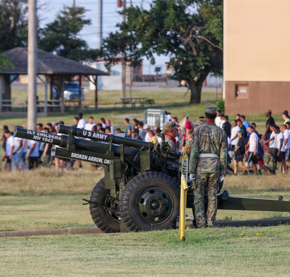 Two U.S. Army soldiers in camouflage uniforms stand beside a ceremonial artillery cannon on a military installation. The cannon bears inscriptions reading 'U.S. ARMY', '2LT CHILDERS MH 1943', and 'BROKEN ARROW, OK'. A large group of people, many in civilian clothing, are gathered in the background, suggesting a military ceremony or demonstration event.