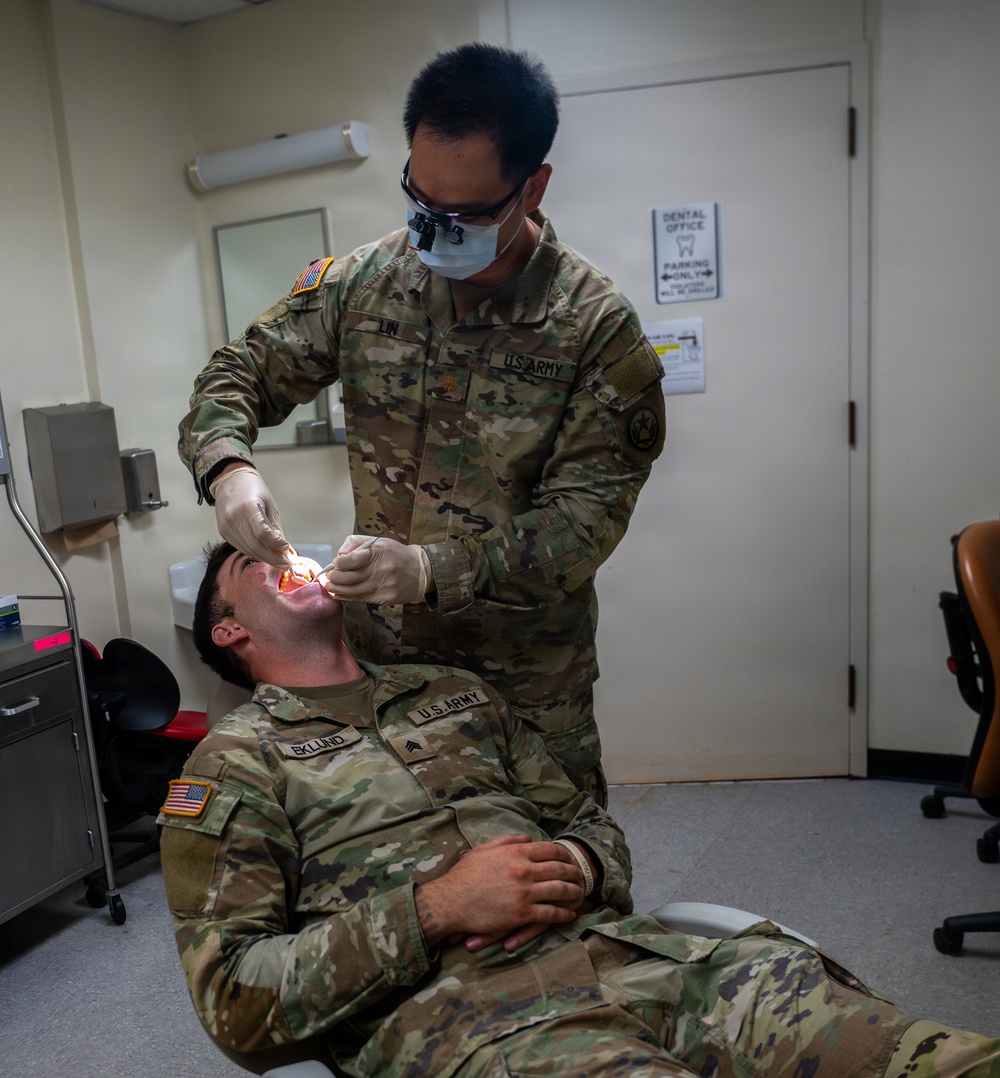 A U.S. Army dental officer with the name tape 'LIN' is performing a dental examination on a fellow soldier identified as 'EKLUND', who is reclined in a dental chair. Both individuals are wearing U.S. Army OCP (Operational Camouflage Pattern) uniforms. The setting is a military dental office, as indicated by a 'Dental Office' sign visible on the door in the background.
