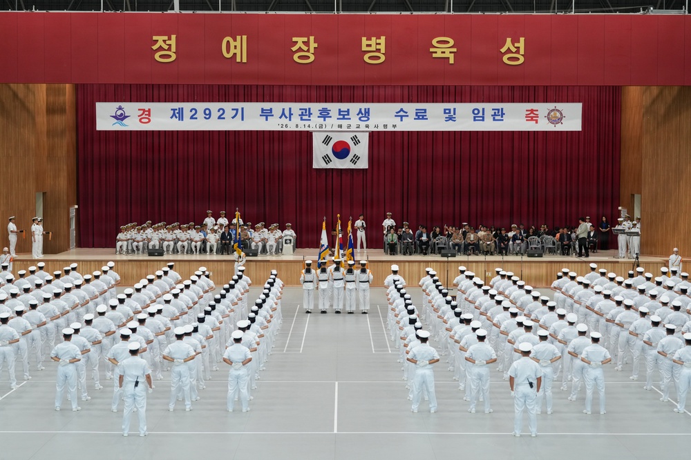 A formal South Korean Navy commissioning ceremony is taking place in a large indoor hall. The banner reads '제292기 부사관후보생 수료 및 임관 축' (292nd Non-Commissioned Officer Candidate Graduation and Commissioning Ceremony), dated 8.14. Rows of naval personnel in white dress uniforms are assembled in formation facing a stage decorated with red curtains and the South Korean flag (Taegukgi).