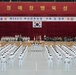 A formal South Korean Navy commissioning ceremony is taking place in a large indoor hall. The banner reads '제292기 부사관후보생 수료 및 임관 축' (292nd Non-Commissioned Officer Candidate Graduation and Commissioning Ceremony), dated 8.14. Rows of naval personnel in white dress uniforms are assembled in formation facing a stage decorated with red curtains and the South Korean flag (Taegukgi).