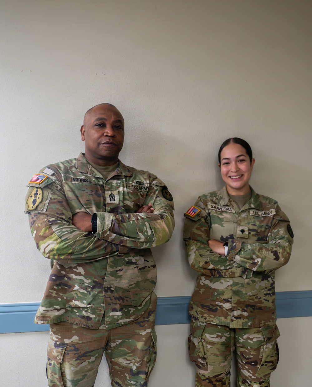 Two U.S. Army soldiers in Operational Camouflage Pattern (OCP) uniforms are standing side by side against an interior wall with arms crossed. The soldier on the left has the name tape 'NORRIS' and displays a senior NCO rank insignia, while the soldier on the right has the name tape 'SAAVEDRA' and is smiling. Both wear U.S. Army branch tapes, American flag patches, and unit patches on their left shoulders.