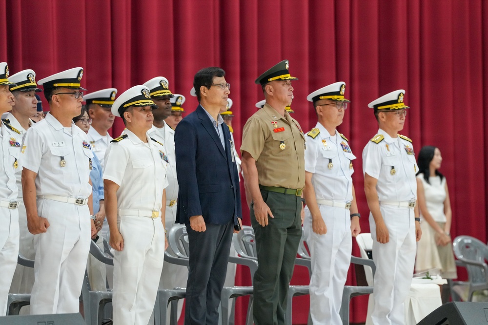 A formal military ceremony featuring personnel in white naval dress uniforms standing at attention on a stage with a red curtain backdrop. The group includes officers from what appears to be multiple nations or branches, along with one civilian in a dark suit and one officer in a tan/khaki uniform. The setting suggests an official ceremony such as a change of command, commissioning, or multinational military event.