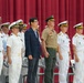 A formal military ceremony featuring personnel in white naval dress uniforms standing at attention on a stage with a red curtain backdrop. The group includes officers from what appears to be multiple nations or branches, along with one civilian in a dark suit and one officer in a tan/khaki uniform. The setting suggests an official ceremony such as a change of command, commissioning, or multinational military event.