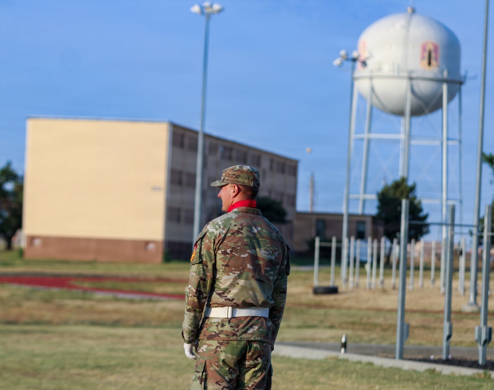 A military service member in US Army Operational Camouflage Pattern (OCP) uniform stands at attention or observation on a military installation parade field. The soldier is wearing ceremonial accessories including a white belt and white gloves, suggesting a formal military ceremony or drill event. A spherical water tower and multi-story barracks building are visible in the background.