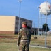 A military service member in US Army Operational Camouflage Pattern (OCP) uniform stands at attention or observation on a military installation parade field. The soldier is wearing ceremonial accessories including a white belt and white gloves, suggesting a formal military ceremony or drill event. A spherical water tower and multi-story barracks building are visible in the background.