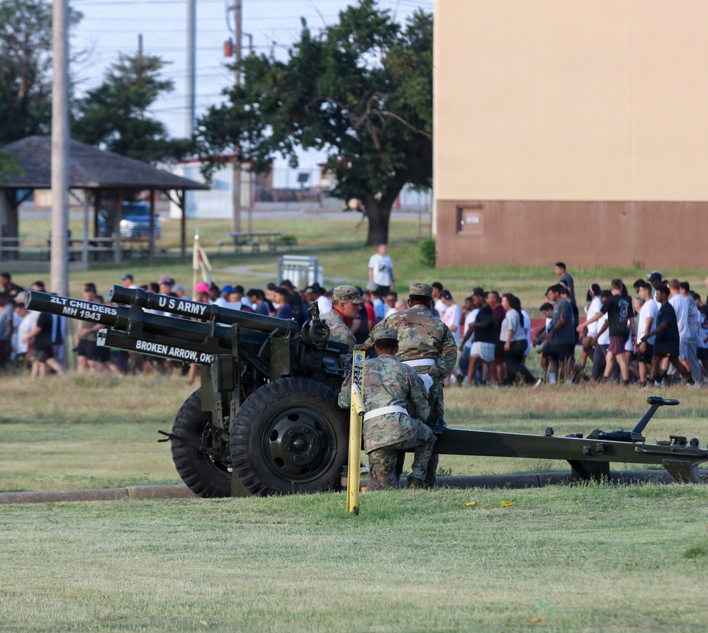 U.S. Army soldiers operate a ceremonial artillery cannon on a military installation, with text on the barrel reading '2LT CHILDERS MH 1943' and 'BROKEN ARROW, OK' indicating a dedication. A large crowd of civilians, likely participants in a community or youth event, can be seen walking in the background. The scene appears to be a military open house, community day, or ceremonial event.