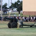 U.S. Army soldiers operate a ceremonial artillery cannon on a military installation, with text on the barrel reading '2LT CHILDERS MH 1943' and 'BROKEN ARROW, OK' indicating a dedication. A large crowd of civilians, likely participants in a community or youth event, can be seen walking in the background. The scene appears to be a military open house, community day, or ceremonial event.