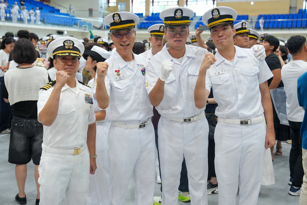 Four individuals in white naval uniforms pose together at what appears to be a military graduation or commissioning ceremony held indoors. They are making fist-pump gestures toward the camera in a celebratory manner. The venue appears to be a large indoor arena or gymnasium filled with other uniformed personnel and civilians.