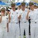 Four individuals in white naval uniforms pose together at what appears to be a military graduation or commissioning ceremony held indoors. They are making fist-pump gestures toward the camera in a celebratory manner. The venue appears to be a large indoor arena or gymnasium filled with other uniformed personnel and civilians.