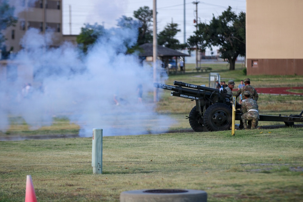 A U.S. Army cannon is being fired on what appears to be a military installation, producing a large cloud of white smoke. Several soldiers in camouflage uniforms are operating the artillery piece, which bears markings identifying it as a U.S. Army cannon from Broken Arrow, Oklahoma. The scene appears to be a ceremonial cannon firing, likely for a military event or retreat ceremony.