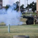 A U.S. Army cannon is being fired on what appears to be a military installation, producing a large cloud of white smoke. Several soldiers in camouflage uniforms are operating the artillery piece, which bears markings identifying it as a U.S. Army cannon from Broken Arrow, Oklahoma. The scene appears to be a ceremonial cannon firing, likely for a military event or retreat ceremony.
