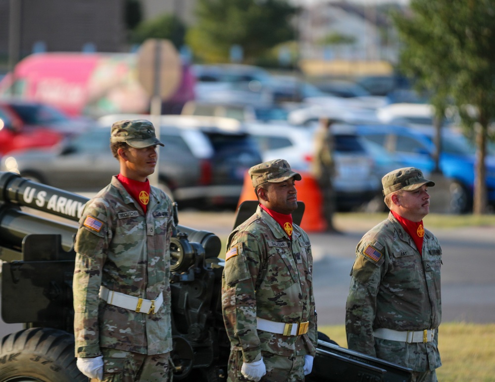Three U.S. Army soldiers in camouflage uniforms stand at attention beside a field artillery cannon marked 'U.S. ARMY' during what appears to be a formal military ceremony. All three wear distinctive red scarves, white gloves, and white belts, indicating a ceremonial detail. The setting appears to be an outdoor military installation with a parking lot visible in the background.