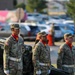 Three U.S. Army soldiers in camouflage uniforms stand at attention beside a field artillery cannon marked 'U.S. ARMY' during what appears to be a formal military ceremony. All three wear distinctive red scarves, white gloves, and white belts, indicating a ceremonial detail. The setting appears to be an outdoor military installation with a parking lot visible in the background.