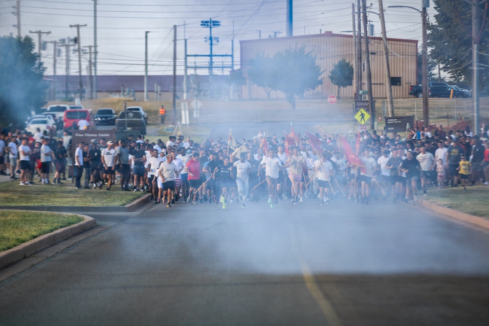A large group of people are participating in an organized run or fun run event on a military installation, evidenced by visible red guidon flags and a sign reading 'Fitness Fitness Center' in the background. Smoke or fog effects are present at the starting line, creating a dramatic atmosphere as hundreds of participants surge forward. The event appears to be a unit physical training run or commemorative run on a military base.