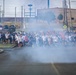 A large group of people are participating in an organized run or fun run event on a military installation, evidenced by visible red guidon flags and a sign reading 'Fitness Fitness Center' in the background. Smoke or fog effects are present at the starting line, creating a dramatic atmosphere as hundreds of participants surge forward. The event appears to be a unit physical training run or commemorative run on a military base.