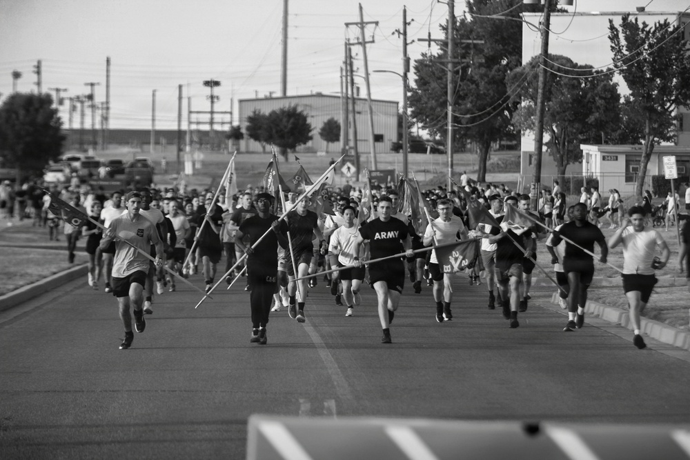 A large group of U.S. Army personnel are conducting a formation run on a paved road, carrying unit guidon flags on poles. The participants are dressed in physical training attire, with at least one person visibly wearing an 'ARMY' shirt. The setting appears to be a military installation, with utility poles, buildings, and trees visible in the background.