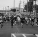 A large group of U.S. Army personnel are conducting a formation run on a paved road, carrying unit guidon flags on poles. The participants are dressed in physical training attire, with at least one person visibly wearing an 'ARMY' shirt. The setting appears to be a military installation, with utility poles, buildings, and trees visible in the background.