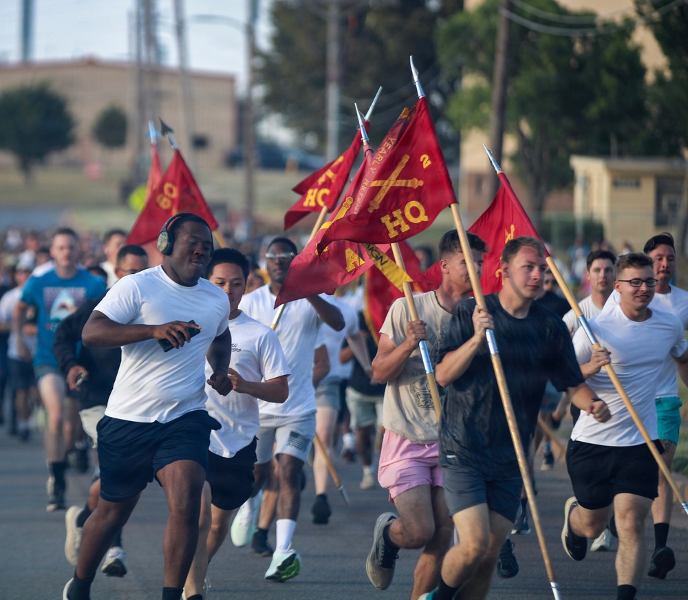 A large group of military personnel in civilian athletic wear are participating in a formation run on a paved road at what appears to be a military installation. Several runners are carrying red guidon flags on wooden poles bearing 'HQ' markings and crossed cannon insignia, consistent with U.S. Marine Corps artillery unit designations. The event appears to be a unit physical training run taking place in warm, sunny conditions.