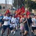 A large group of military personnel in civilian athletic wear are participating in a formation run on a paved road at what appears to be a military installation. Several runners are carrying red guidon flags on wooden poles bearing 'HQ' markings and crossed cannon insignia, consistent with U.S. Marine Corps artillery unit designations. The event appears to be a unit physical training run taking place in warm, sunny conditions.