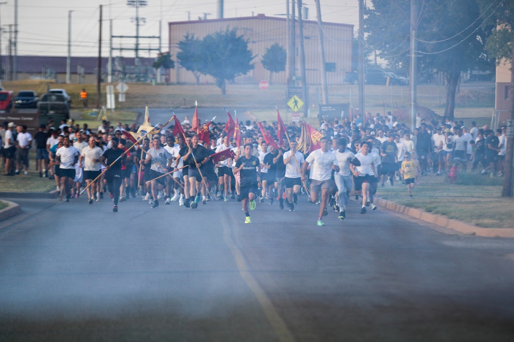 A large group of runners, likely military personnel, are charging down a road carrying unit guidon flags and pennants on staffs. The scene appears to be a formation run or physical training event on a military installation, with smoke or haze visible in the foreground. Hundreds of participants dressed in athletic wear — predominantly white and black shirts — fill the road.