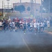 A large group of runners, likely military personnel, are charging down a road carrying unit guidon flags and pennants on staffs. The scene appears to be a formation run or physical training event on a military installation, with smoke or haze visible in the foreground. Hundreds of participants dressed in athletic wear — predominantly white and black shirts — fill the road.