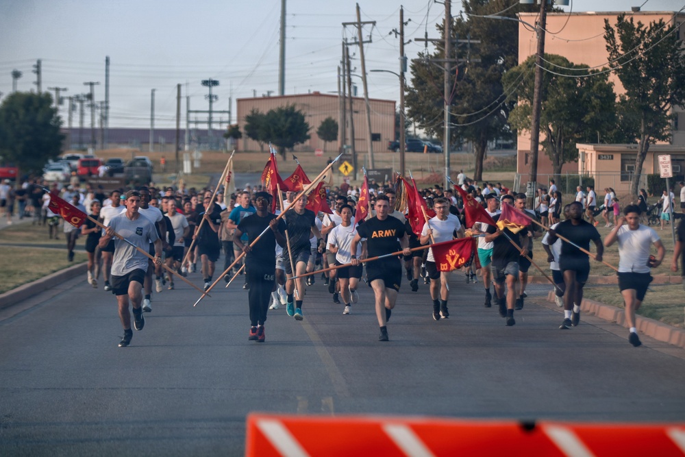 A large group of military personnel and civilians are participating in a formation run on a paved road at what appears to be a U.S. Army installation. Several runners in the front carry unit guidon flags (pennants on wooden poles) with red and gold coloring, consistent with U.S. Army unit colors. The event appears to be an organized physical training or morale run, likely on a military base.