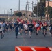 A large group of military personnel and civilians are participating in a formation run on a paved road at what appears to be a U.S. Army installation. Several runners in the front carry unit guidon flags (pennants on wooden poles) with red and gold coloring, consistent with U.S. Army unit colors. The event appears to be an organized physical training or morale run, likely on a military base.
