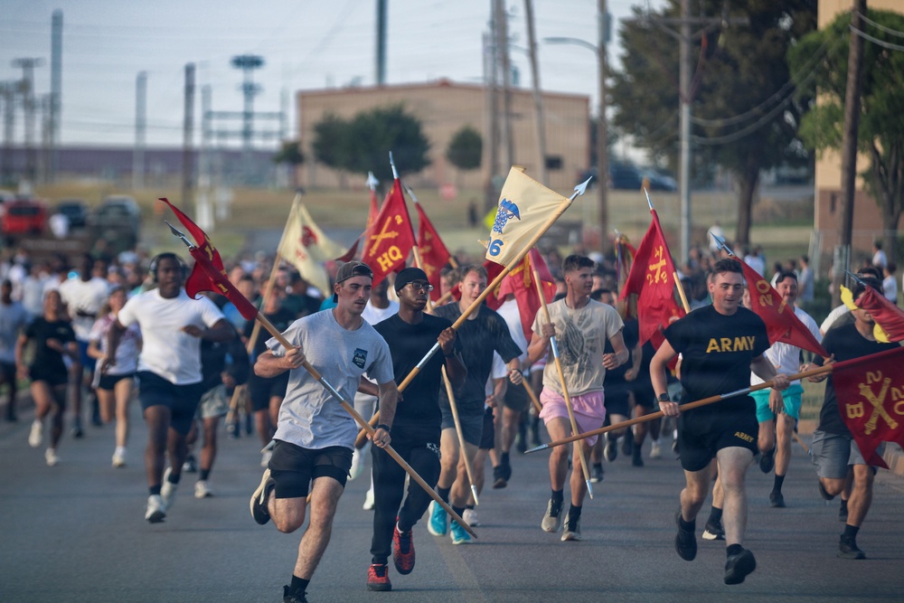 A large group of people, including U.S. Army personnel in PT gear, are running down a road while carrying military guidon flags. The flags are red and tan/cream colored, bearing unit designations including 'HQ' and numerical identifiers. The event appears to be a unit formation run or military physical training exercise on a military installation.