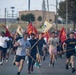 A large group of people, including U.S. Army personnel in PT gear, are running down a road while carrying military guidon flags. The flags are red and tan/cream colored, bearing unit designations including 'HQ' and numerical identifiers. The event appears to be a unit formation run or military physical training exercise on a military installation.