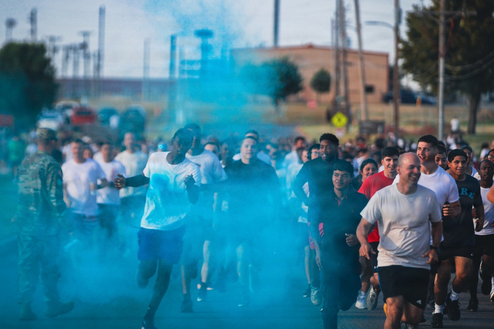 A large group of people are participating in what appears to be a color run or fun run event, running through a cloud of bright teal/cyan colored powder or smoke. The event appears to take place on a road or street near a military or institutional facility, with utility poles, a brick building, and trees visible in the background. Participants are wearing casual athletic clothing, predominantly white t-shirts and shorts.