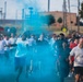 A large group of people are participating in what appears to be a color run or fun run event, running through a cloud of bright teal/cyan colored powder or smoke. The event appears to take place on a road or street near a military or institutional facility, with utility poles, a brick building, and trees visible in the background. Participants are wearing casual athletic clothing, predominantly white t-shirts and shorts.