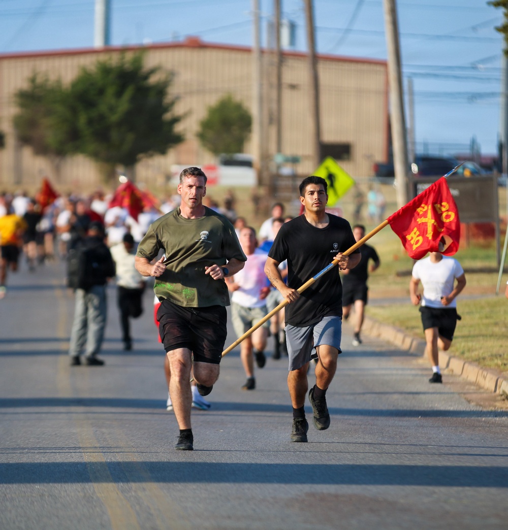 A group of runners participating in what appears to be a military formation run or commemorative race on a road. Two lead runners are prominently visible in the foreground, with one carrying a red military-style guidon flag on a wooden pole. A large crowd of participants follows behind them on the road.