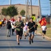 A group of runners participating in what appears to be a military formation run or commemorative race on a road. Two lead runners are prominently visible in the foreground, with one carrying a red military-style guidon flag on a wooden pole. A large crowd of participants follows behind them on the road.
