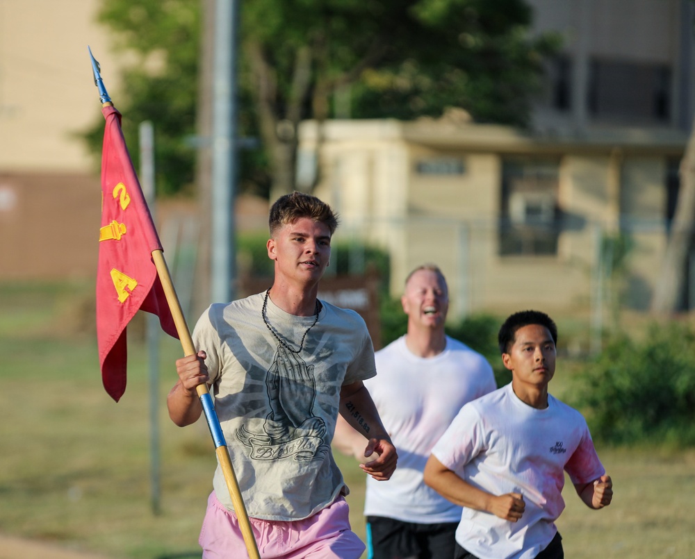 Three individuals are running outdoors, with the lead runner carrying a red military-style guidon flag with yellow markings, including what appears to be the number '2' and letter 'A'. The setting appears to be a military base or institutional campus based on the buildings and grounds visible in the background. The lead runner wears a graphic t-shirt and pink shorts, while the two runners behind him wear white t-shirts.