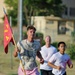 Three individuals are running outdoors, with the lead runner carrying a red military-style guidon flag with yellow markings, including what appears to be the number '2' and letter 'A'. The setting appears to be a military base or institutional campus based on the buildings and grounds visible in the background. The lead runner wears a graphic t-shirt and pink shorts, while the two runners behind him wear white t-shirts.
